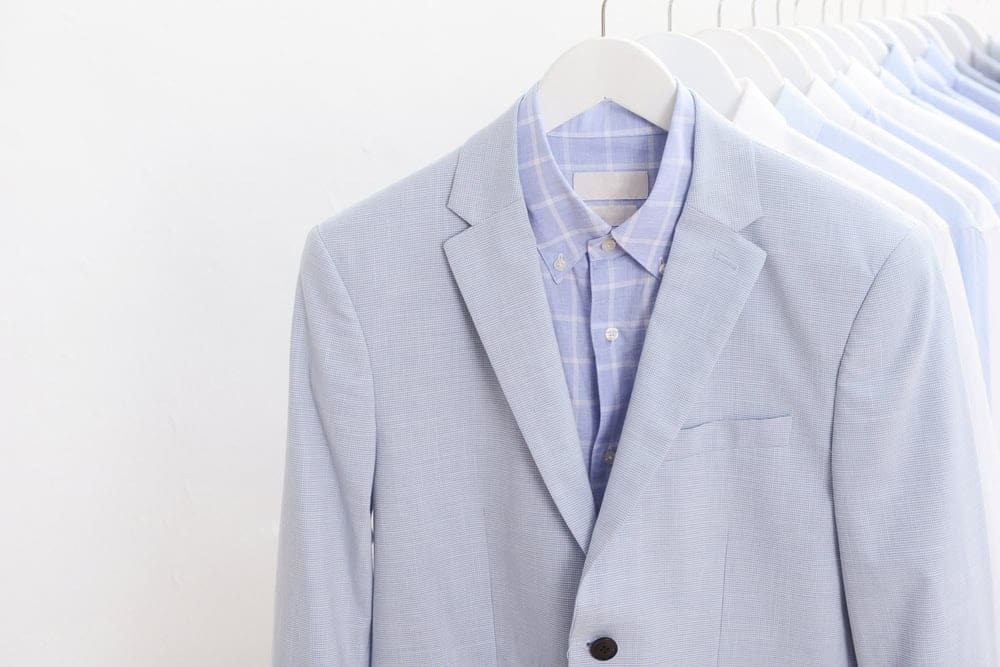 A light gray suit jacket over a blue plaid dress shirt hangs on a rack with several other light-colored shirts in the background.