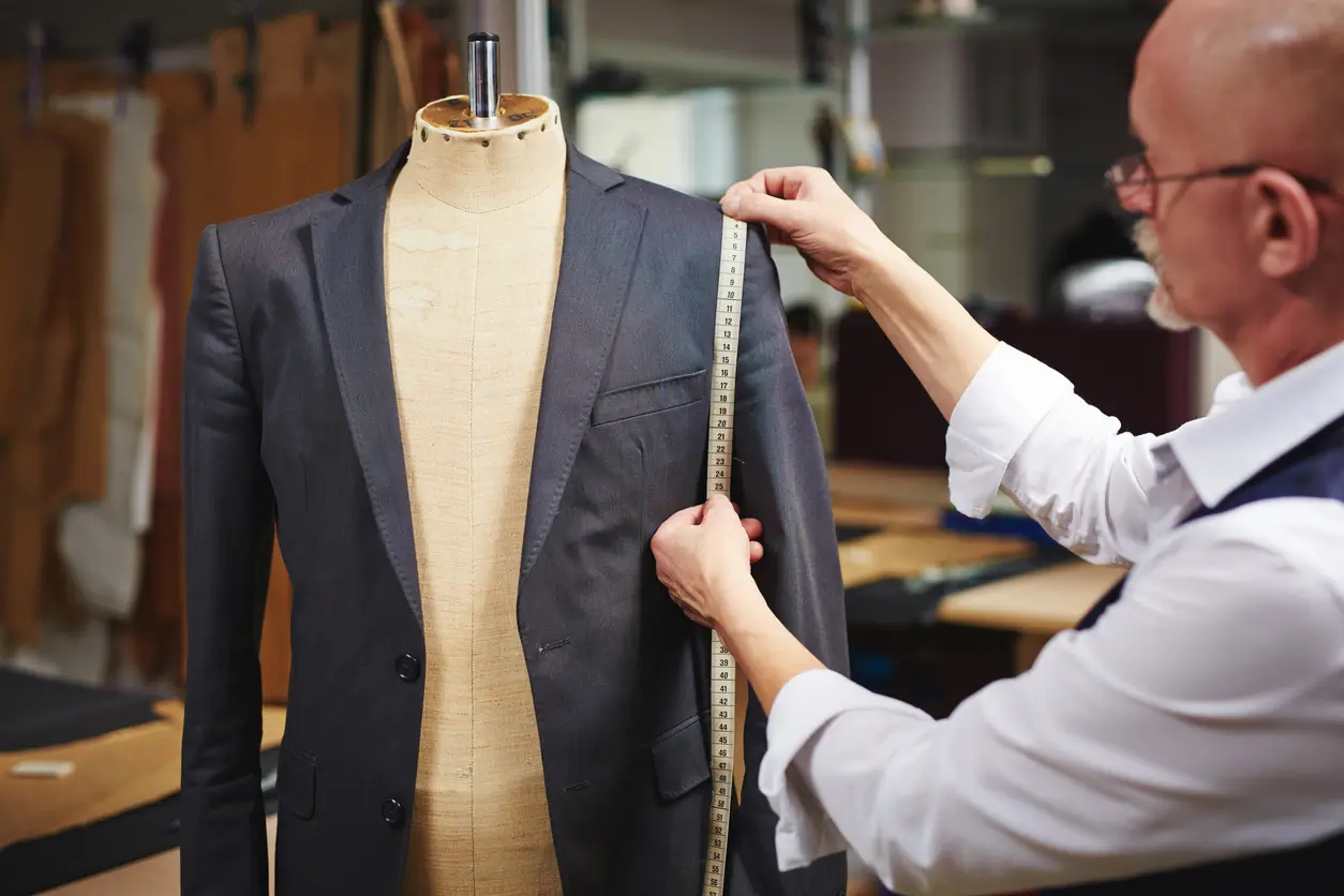 Tailor measuring the shoulder of a suit jacket on a dress form with a measuring tape in a workshop.